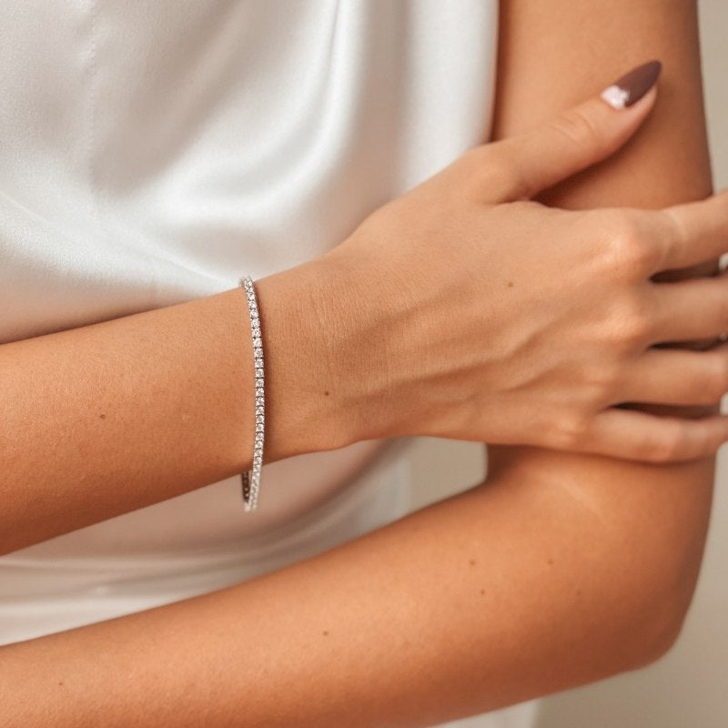 Gros plan sur le bras d'une femme portant un bracelet tennis en argent avec des pierres de zirconia, sur un fond de satin blanc.