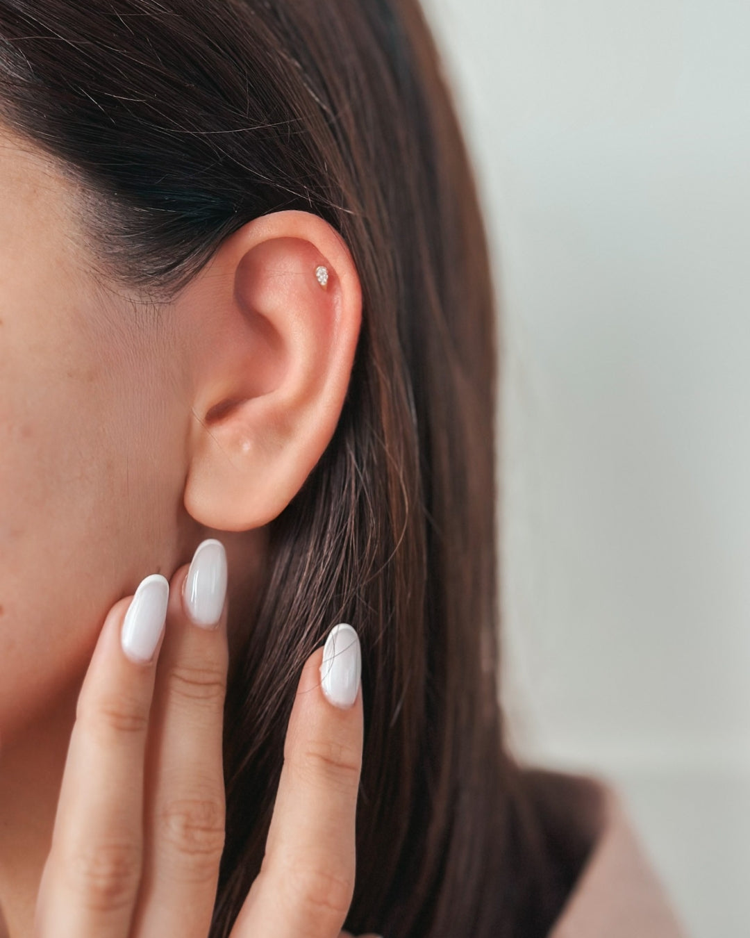 Gros plan sur une oreille avec une petite boucle d'oreille clou et des doigts avec des ongles vernis en blanc.
