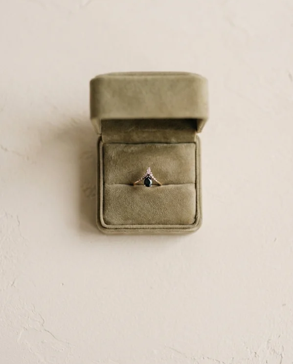 Une boîte à bijoux en velours vert ouverte contenant une bague avec une pierre foncée, présentée sur une surface texturée claire.
