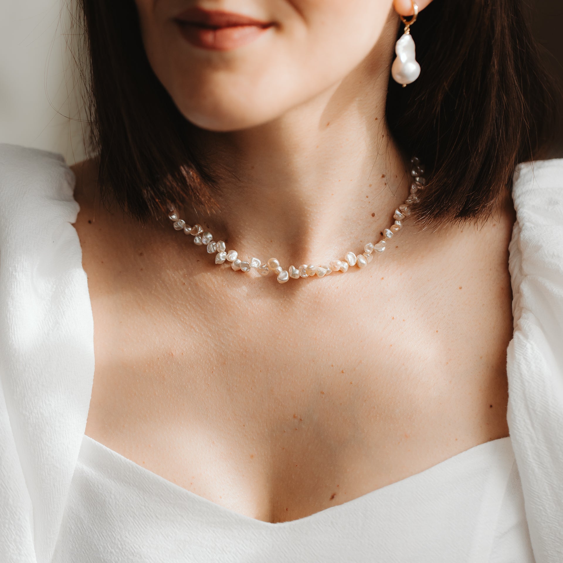 Gros plan d’un mannequin portant un délicat collier en perles keshi roses et une boucle d’oreille pendante en perle baroque sur monture plaquée or, avec une robe blanche sous une douce lumière naturelle.
