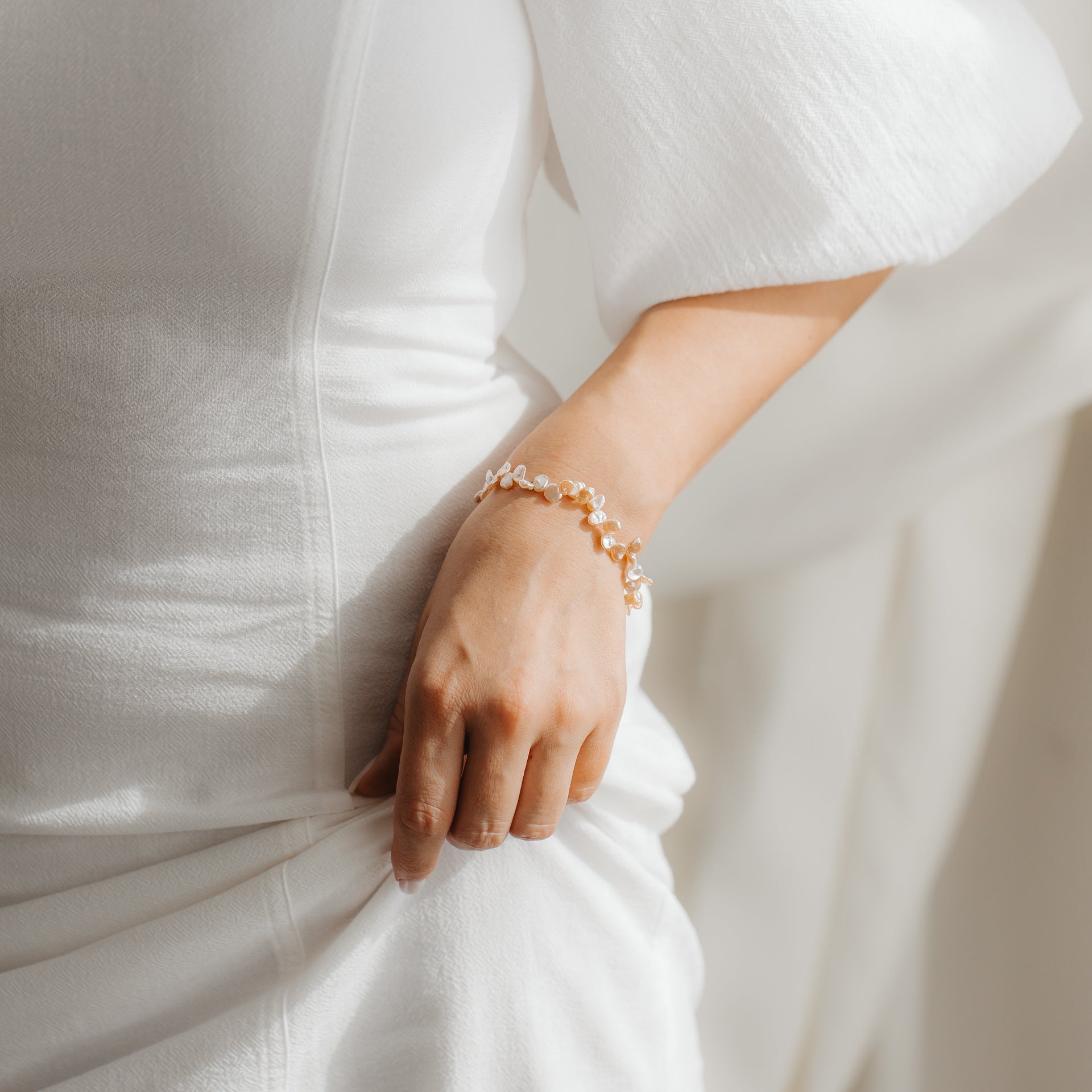 Gros plan d'un bracelet en argent sterling plaqué or avec des perles keshi roses au poignet d'une personne, mis en valeur sur une robe blanche sous une lumière douce.