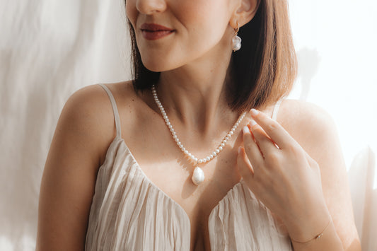 Gros plan d’une femme portant un pendentif en perle baroque sur un collier de perles, avec une boucle d’oreille pendante en perle assortie en argent plaqué or, éclairée par une douce lumière naturelle.