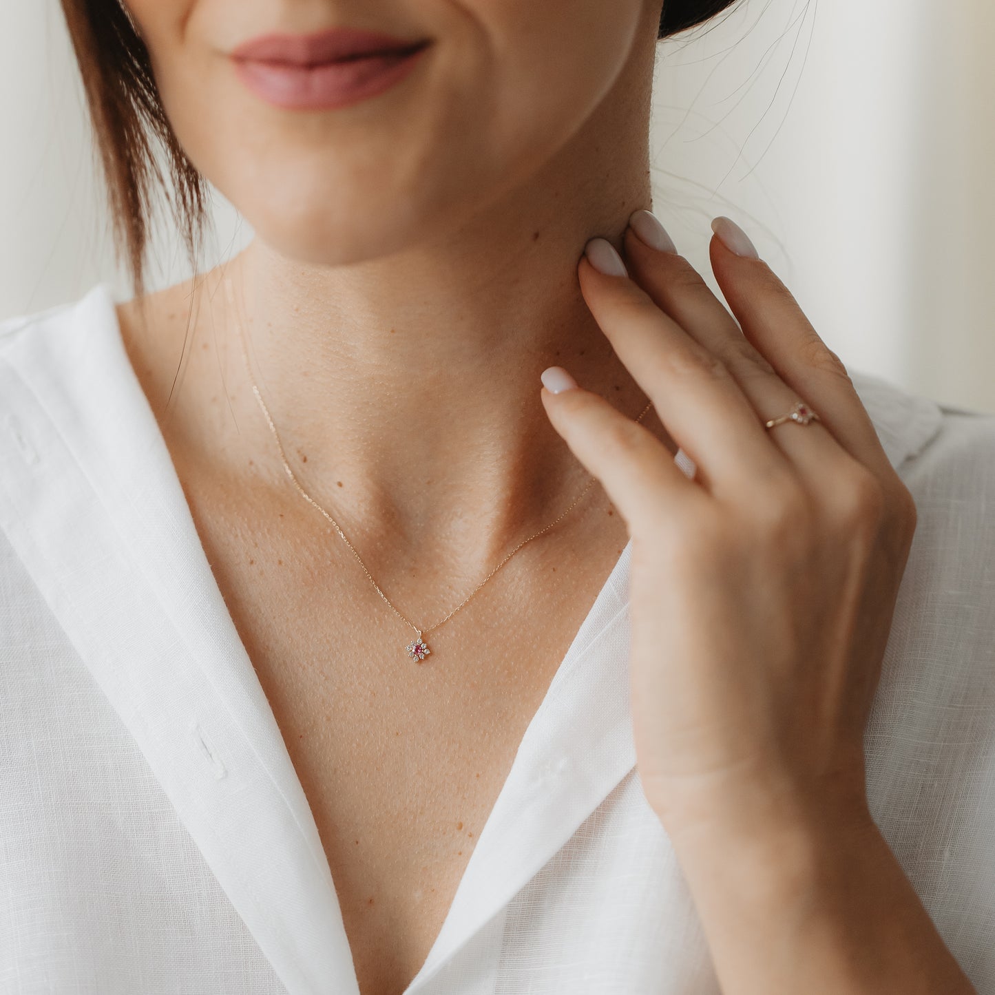 Gros plan d’une personne portant le collier Flora en or 14k, avec un petit pendentif en forme de fleur serti de tourmaline rose et de saphirs blancs, la main effleurant le cou tout en portant un chemisier blanc.