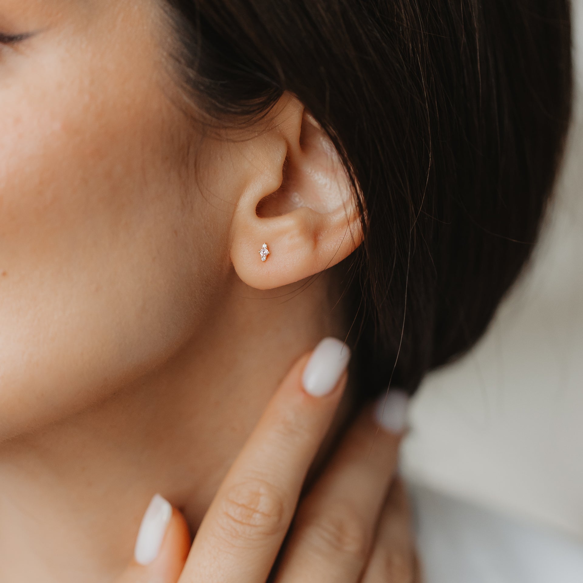 Gros plan d’une oreille portant une petite puce d’oreille Flora en or jaune 14K avec morganite rose et saphir blanc, tandis qu’une main à la manucure neutre touche le cou.