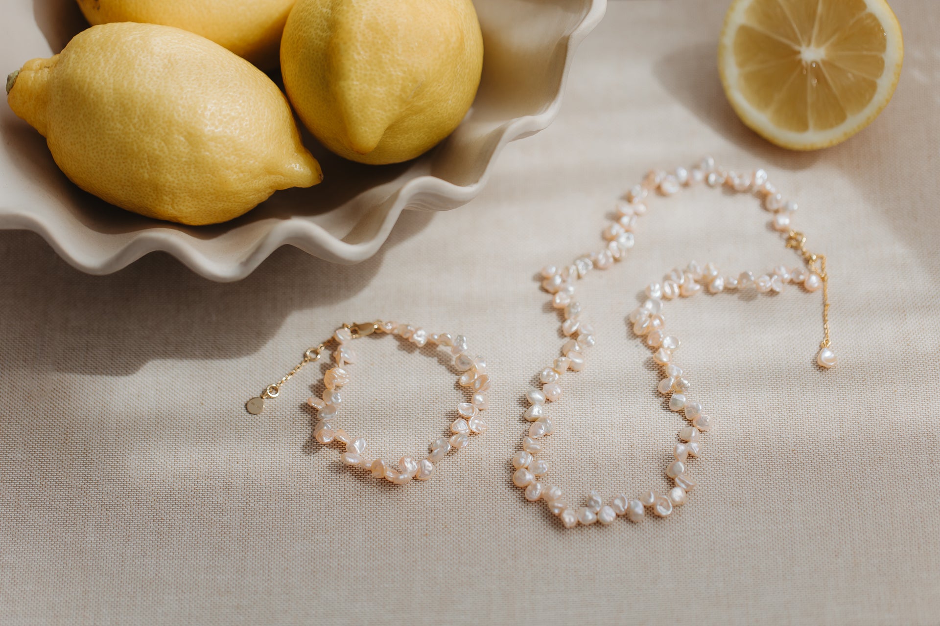 Collier en perles keshi roses et bracelet assorti en argent plaqué or, présentés sur du lin beige avec des citrons frais.