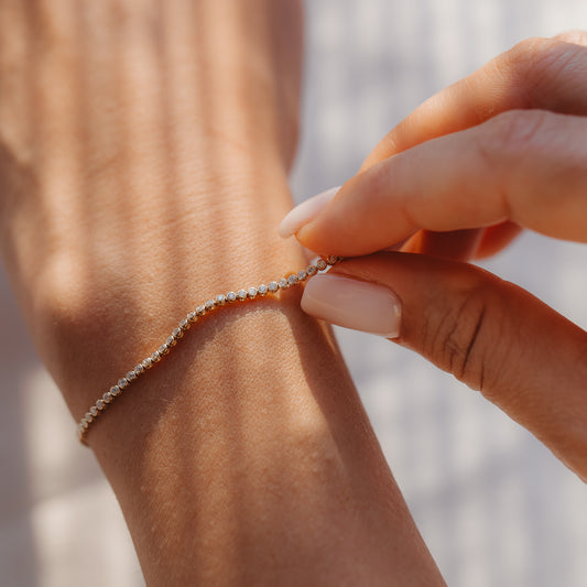 Gros plan d'une main qui attache un bracelet tennis en diamants en or jaune 14K autour d'un poignet, à la lumière chaude du soleil.