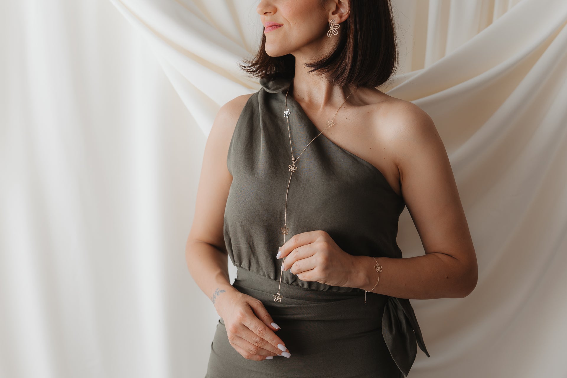 Gros plan d’un mannequin portant le collier lariat Wildflower Maxi en or jaune 14K, orné de délicates breloques en forme de fleur, superposé à une robe vert olive à une épaule, devant un fond crème drapé et doux.