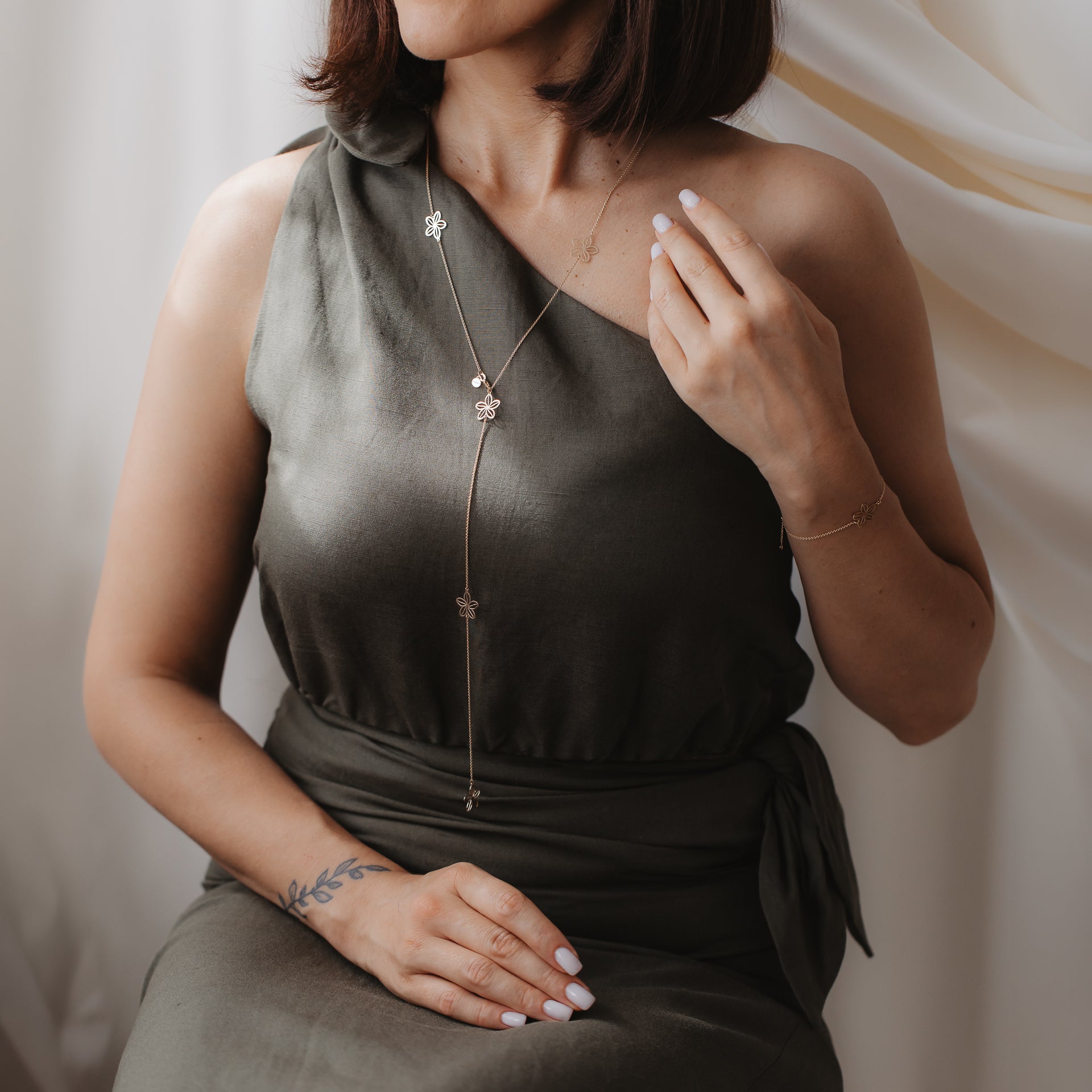 Gros plan d’un mannequin portant le collier lariat Wildflower Maxi en or jaune 14K avec des breloques florales, sur une robe à une épaule couleur olive.