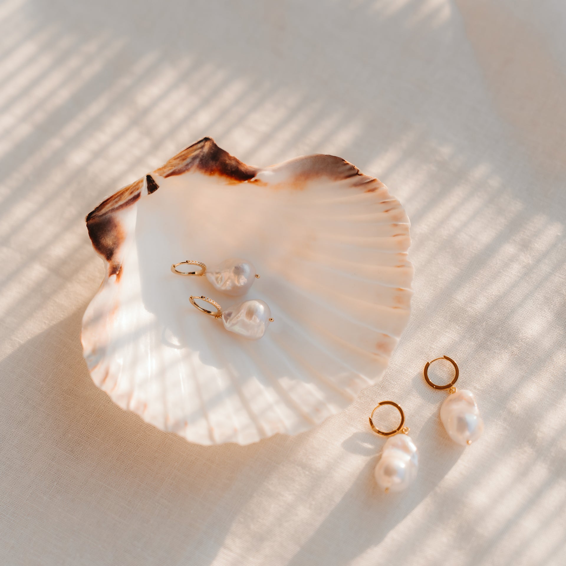 Élégantes boucles d’oreilles pendantes à perles baroques, créoles en argent sterling plaqué or rehaussées de détails en oxyde de zirconium, présentées sur une coquille Saint‑Jacques posée sur du lin sous une lumière du jour chaleureuse.
