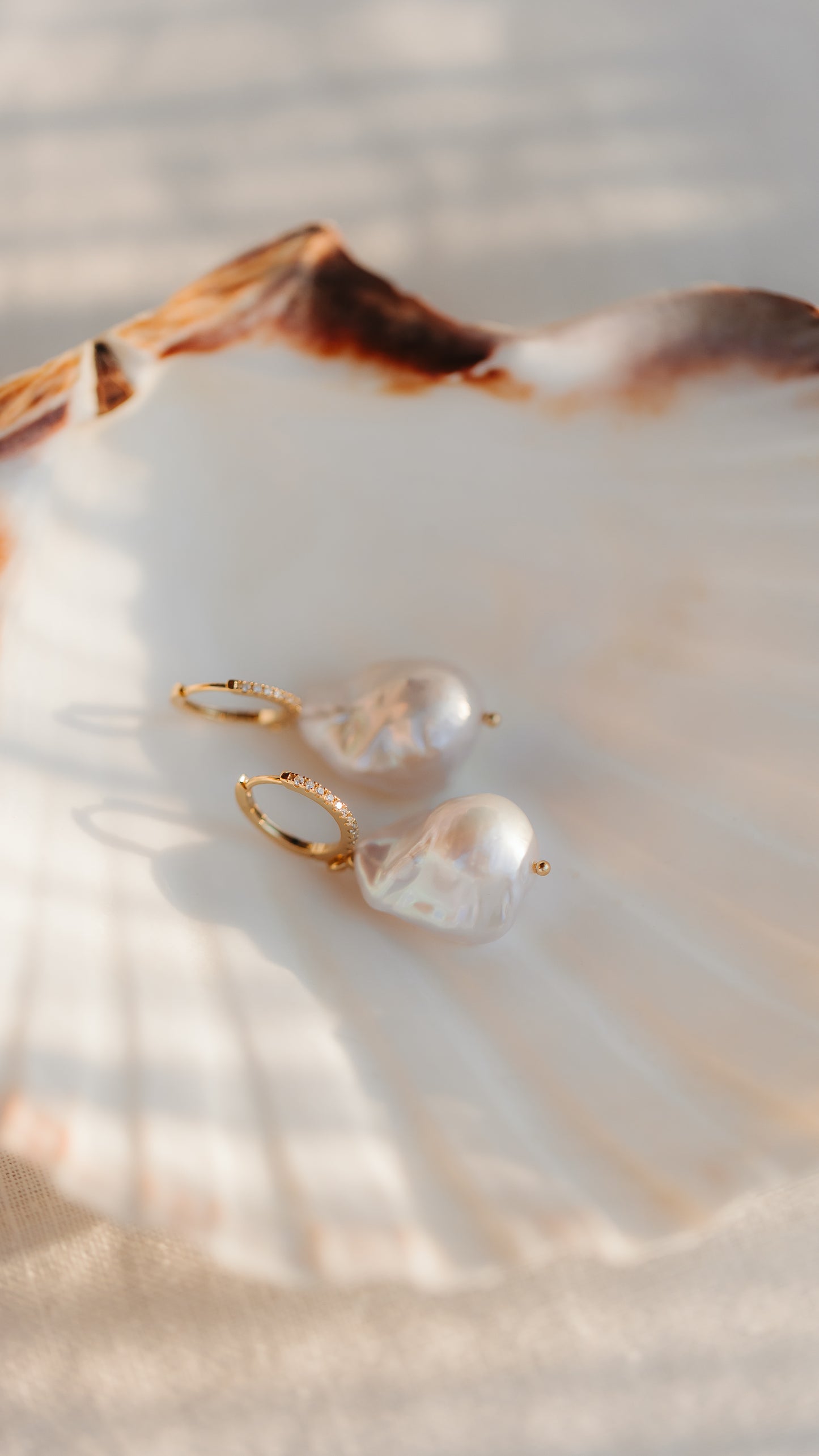 Boucles d’oreilles créoles en argent 925 plaqué or avec de grandes perles baroques, mises en scène sur une coquille Saint‑Jacques sous une lumière naturelle chaleureuse.