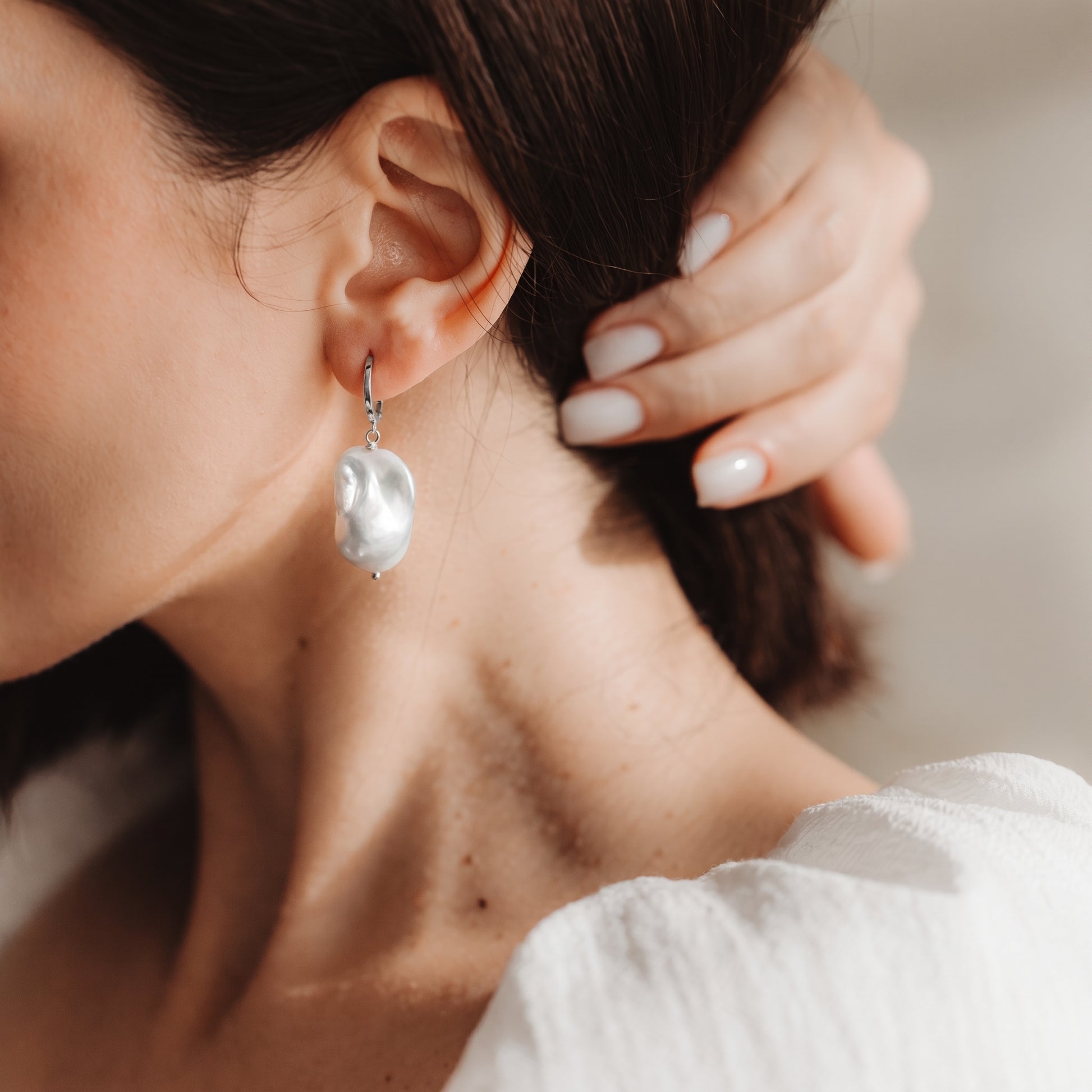 Gros plan d’une oreille portant une créole en argent 925 plaqué rhodium, ornée d’une grande perle baroque pendante, tandis qu’une main touche les cheveux.