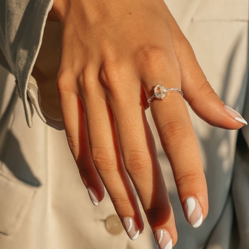 Gros plan d'une main portant une bague en argent avec une pierre de lune naturelle sur fond beige.