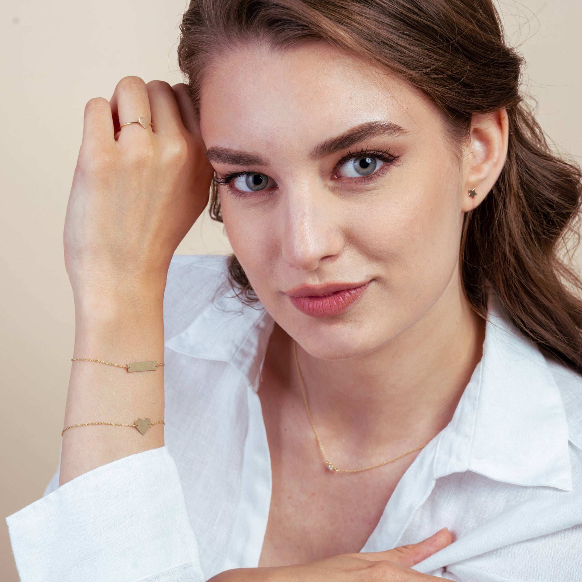 Femme portant une chemise blanche, présentant des bijoux en or incluant un bracelet cœur, des petites boucles d'oreilles et un collier délicat dans un studio.