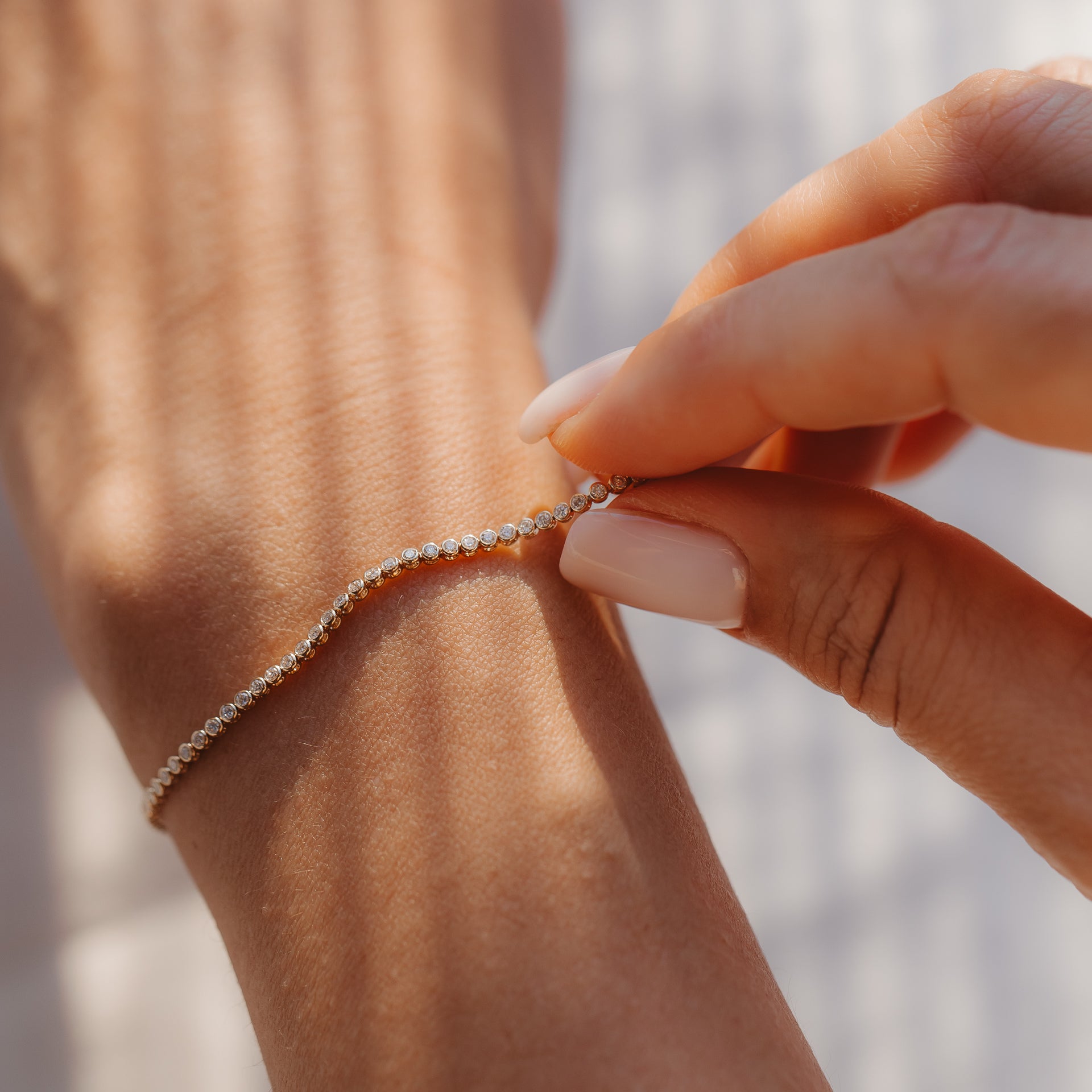 Gros plan d'une main attachant un bracelet tennis en or jaune 14K serti de diamants au poignet, sous une douce lumière du soleil.