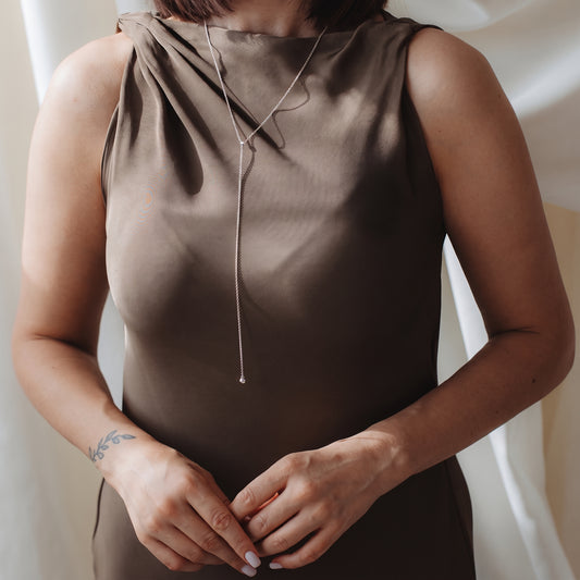 Gros plan d’une personne portant un collier bola (lariat) en argent sterling sur une robe en satin taupe, dans une douce lumière intérieure.