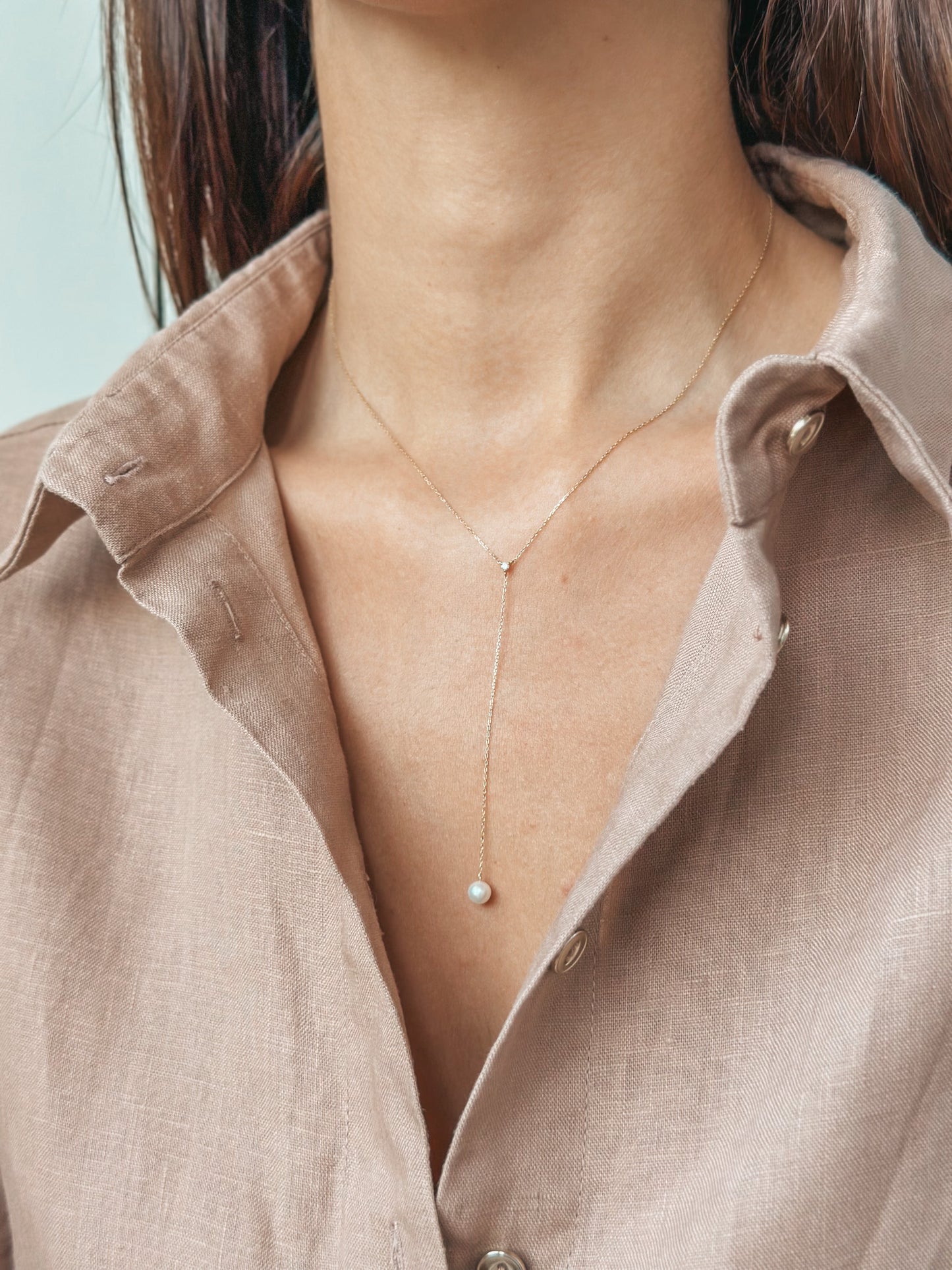 Une femme portant un collier en or avec un petit pendentif en perle, chemise beige partiellement visible