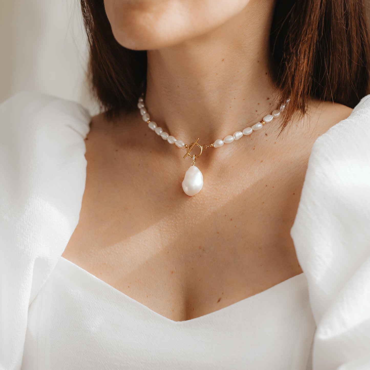 Gros plan d'une femme portant un collier court de perles naturelles, orné d'un pendentif en perle baroque et d'un fermoir toggle en or, assorti à une robe blanche.