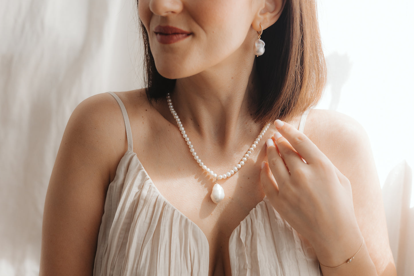 Gros plan d’une femme portant un pendentif en perle baroque sur un collier de perles, avec une boucle d’oreille pendante en perle assortie en argent plaqué or, éclairée par une douce lumière naturelle.
