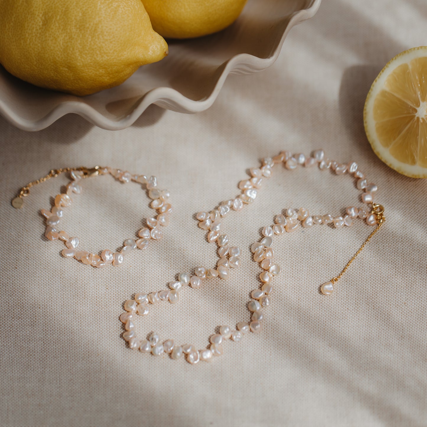 Collier en argent plaqué or avec des perles keshi roses, présenté sur du lin neutre avec des citrons, avec un bracelet assorti à proximité.
