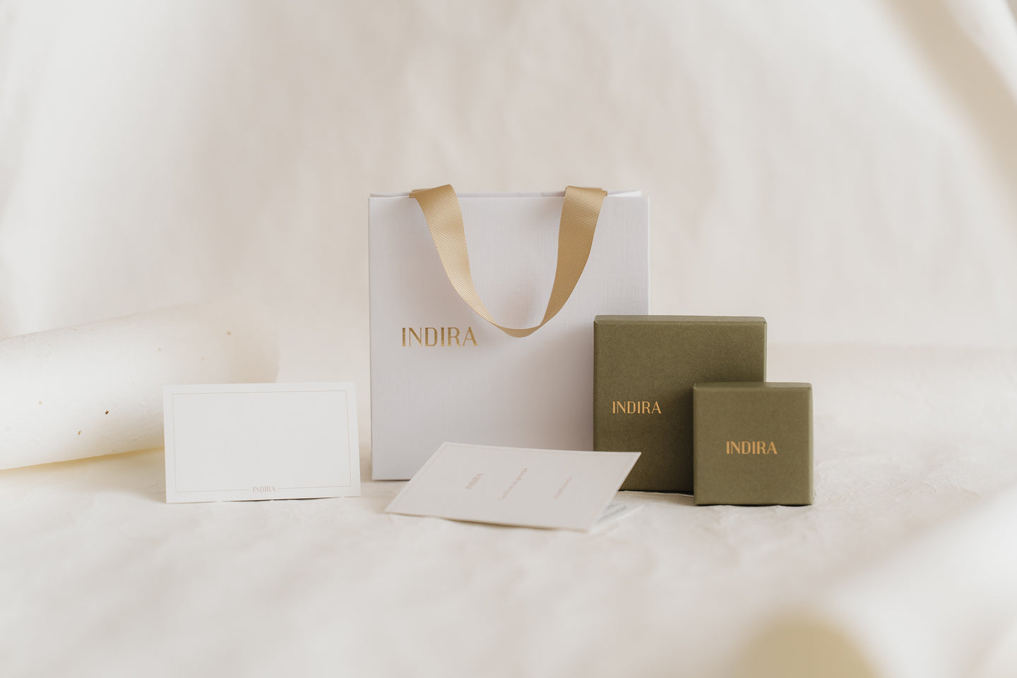 Photo de studio minimaliste de l'ensemble d'emballage cadeau INDIRA : un sac blanc avec anses en ruban doré, deux boîtes à bijoux vert olive et des cartes vierges sur un fond ivoire doux.