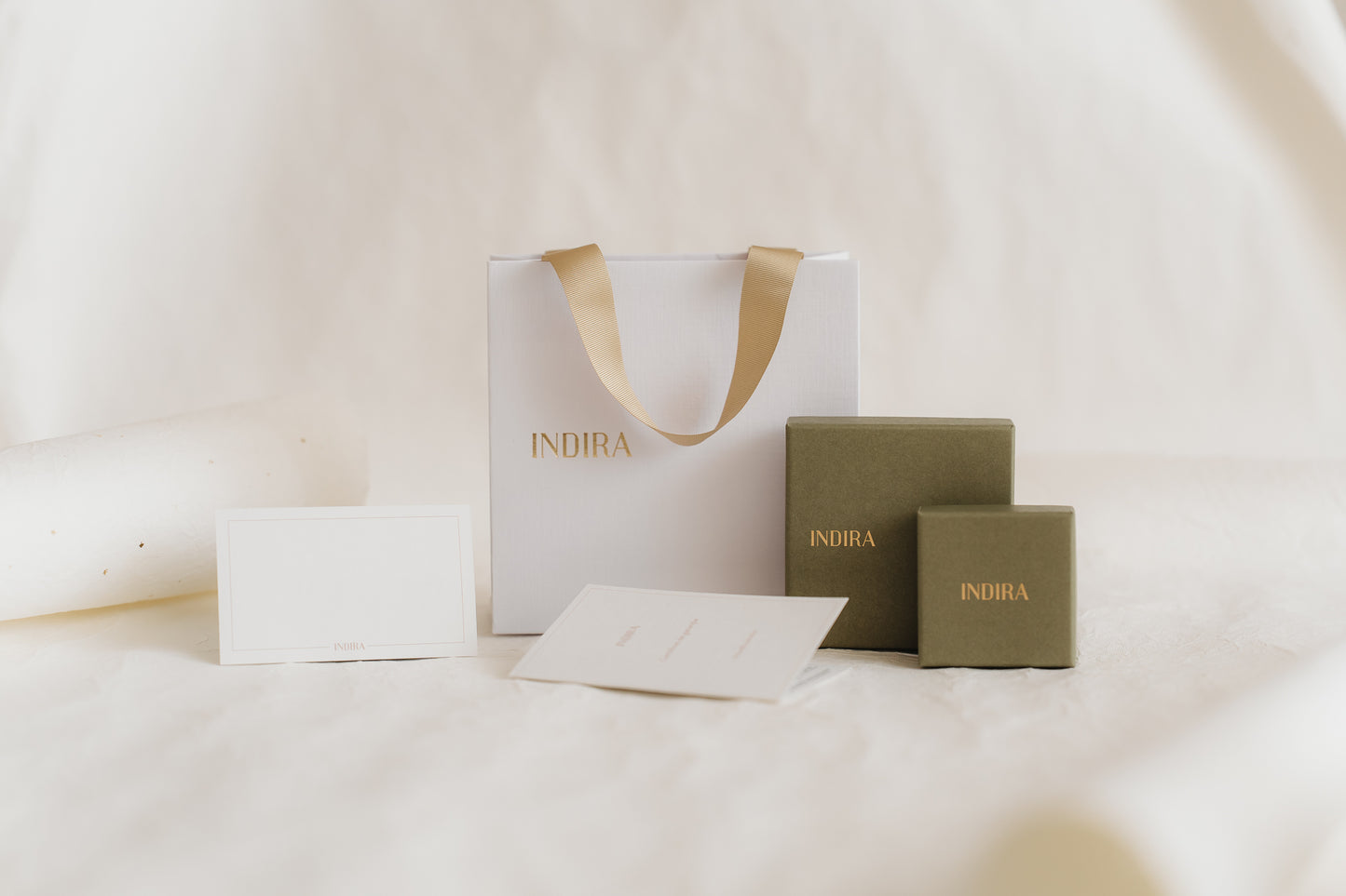 Photo de studio minimaliste de l'ensemble d'emballage cadeau INDIRA : un sac blanc avec anses en ruban doré, deux boîtes à bijoux vert olive et des cartes vierges sur un fond ivoire doux.