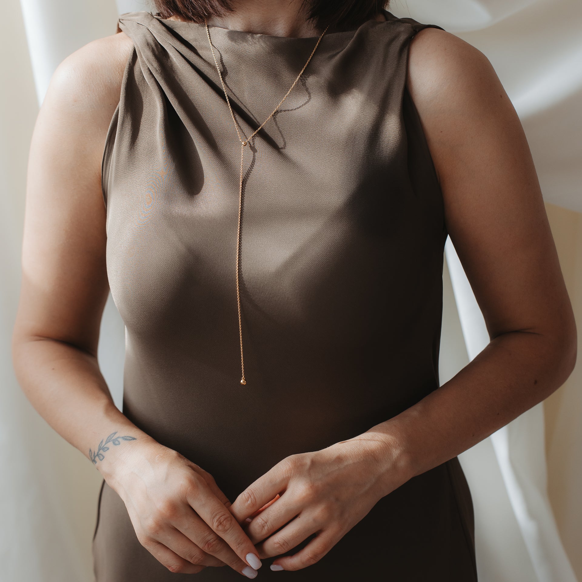 Gros plan d’une personne portant un collier lariat bola en argent 925 plaqué or sur une robe en satin marron, les mains délicatement jointes.