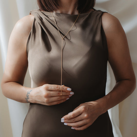 Gros plan d’une personne portant un collier lariat (Bola) en argent 925 plaqué or sur une robe en satin taupe, ses mains ajustant délicatement le pendentif goutte.