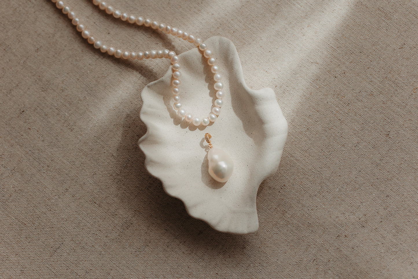 Pendentif en perle baroque avec bélière en argent plaqué or, posé dans une coupelle en céramique en forme de coquillage aux bords festonnés, avec un rang de perles sur un fond de lin beige.