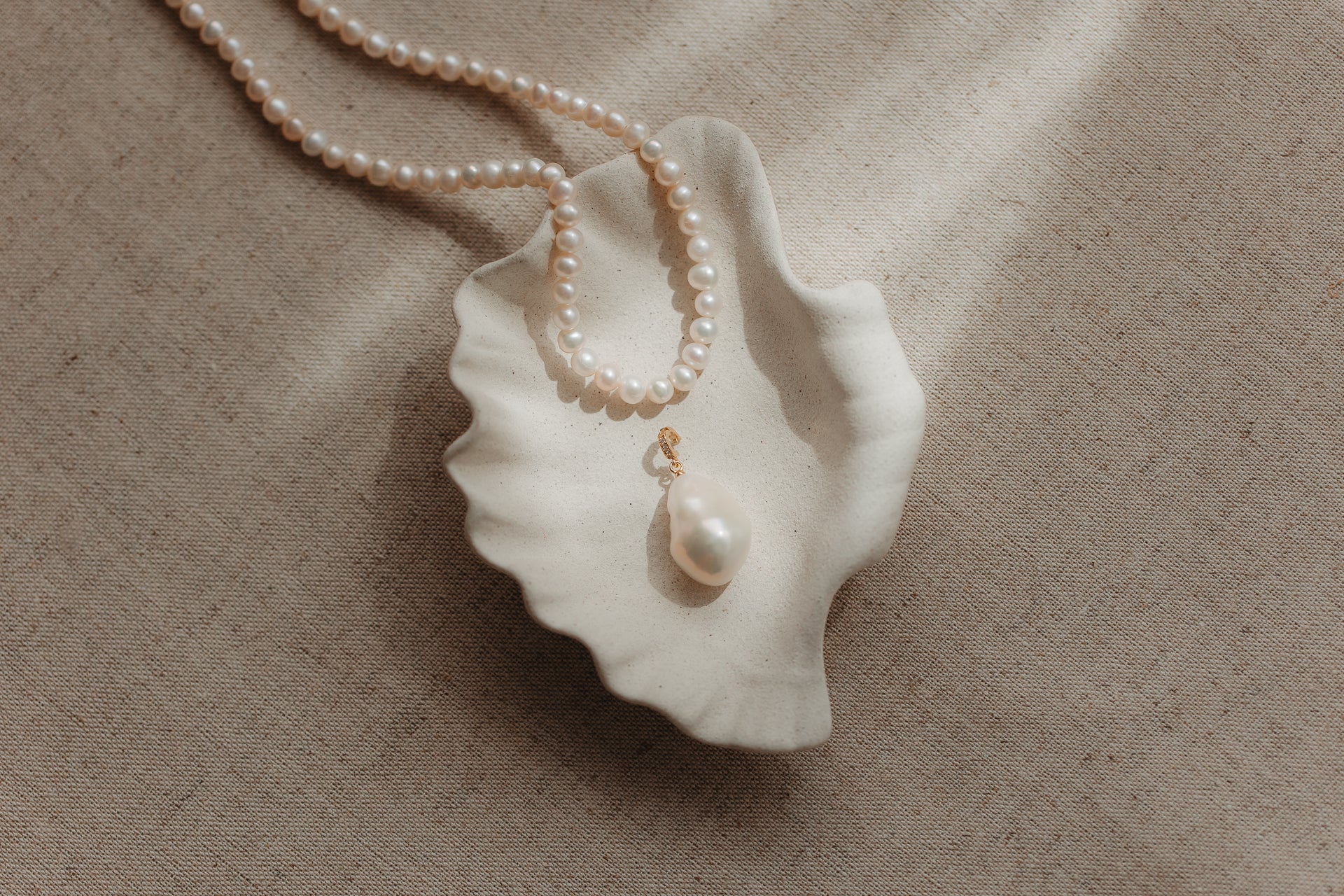 Pendentif en perle baroque avec bélière en argent plaqué or, posé dans une coupelle en céramique en forme de coquillage aux bords festonnés, avec un rang de perles sur un fond de lin beige.