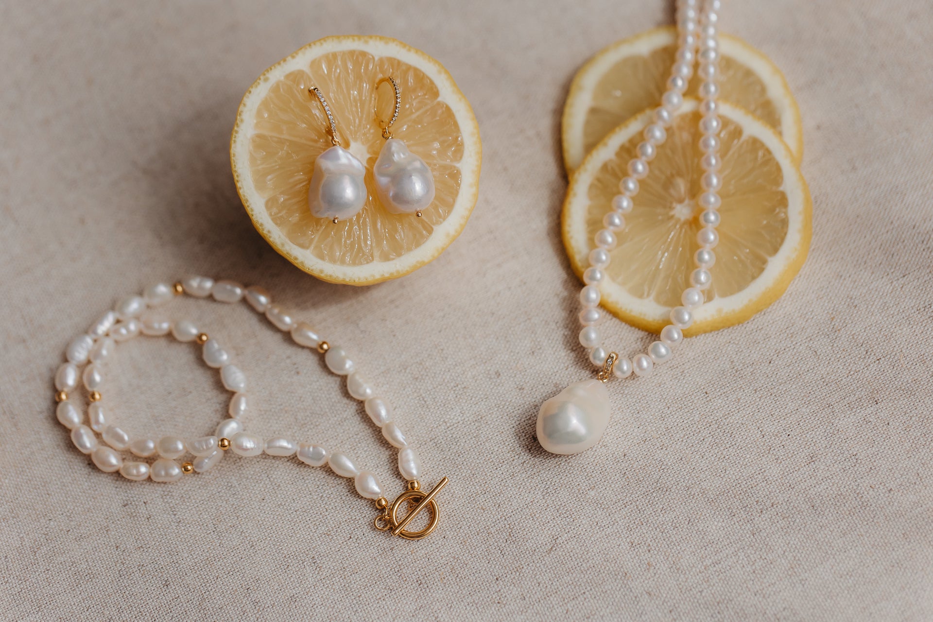 Boucles d’oreilles en perles baroques sur des créoles en argent sterling plaqué or, piquées dans une moitié de citron, avec collier et bracelet en perles assortis présentés sur du lin beige.