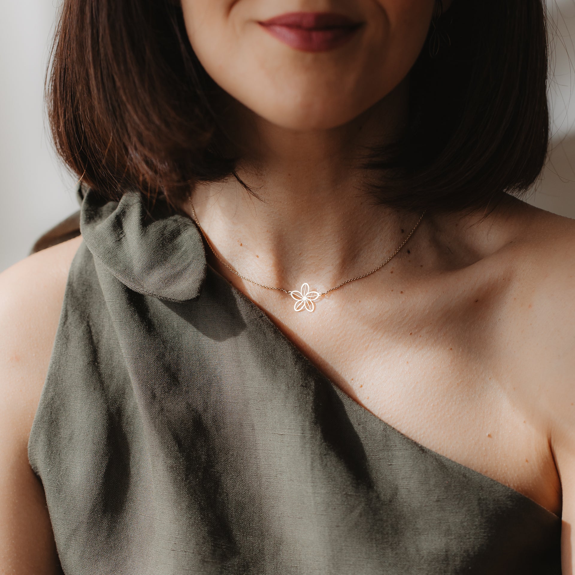 Gros plan d’une personne portant un collier délicat en or jaune 14K présentant le contour d’une fleur sauvage au niveau de la clavicule, assorti à un top olive asymétrique à une épaule, en lumière naturelle.