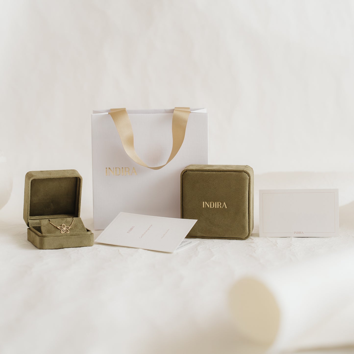 Élégant ensemble d’emballage cadeau Indira avec un sac blanc aux anses en ruban doré, des écrins en velours olive (dont un ouvert) et des cartes, disposés sur un fond doux et neutre.