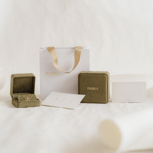 Élégant ensemble d’emballage cadeau Indira avec un sac blanc aux anses en ruban doré, des écrins en velours olive (dont un ouvert) et des cartes, disposés sur un fond doux et neutre.