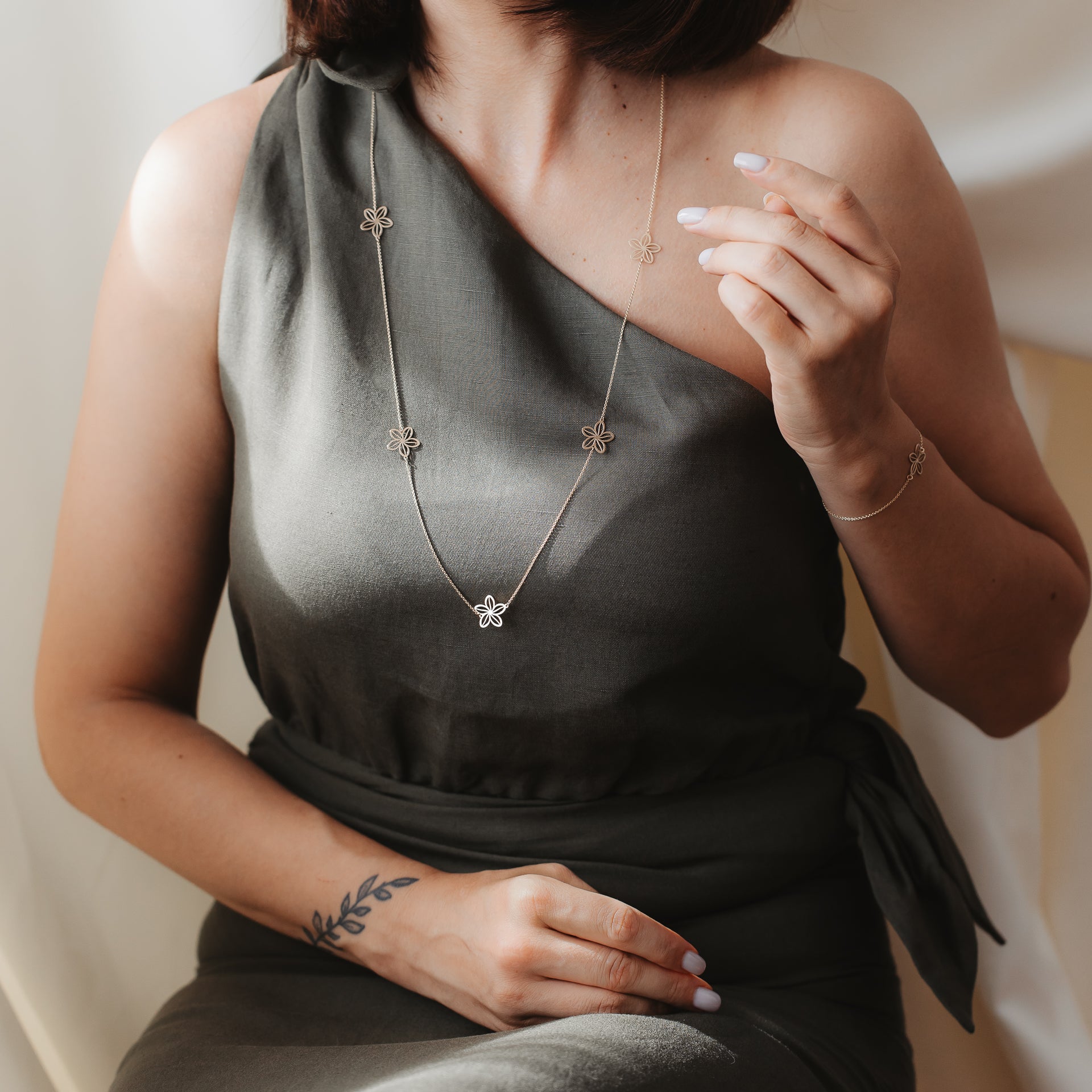 Gros plan d’un mannequin en robe vert olive à une épaule, portant le collier à stations Wildflower Maxi en or jaune 14K avec de petites breloques en forme de fleur, photographié sous une lumière intérieure douce avec un bracelet assorti.