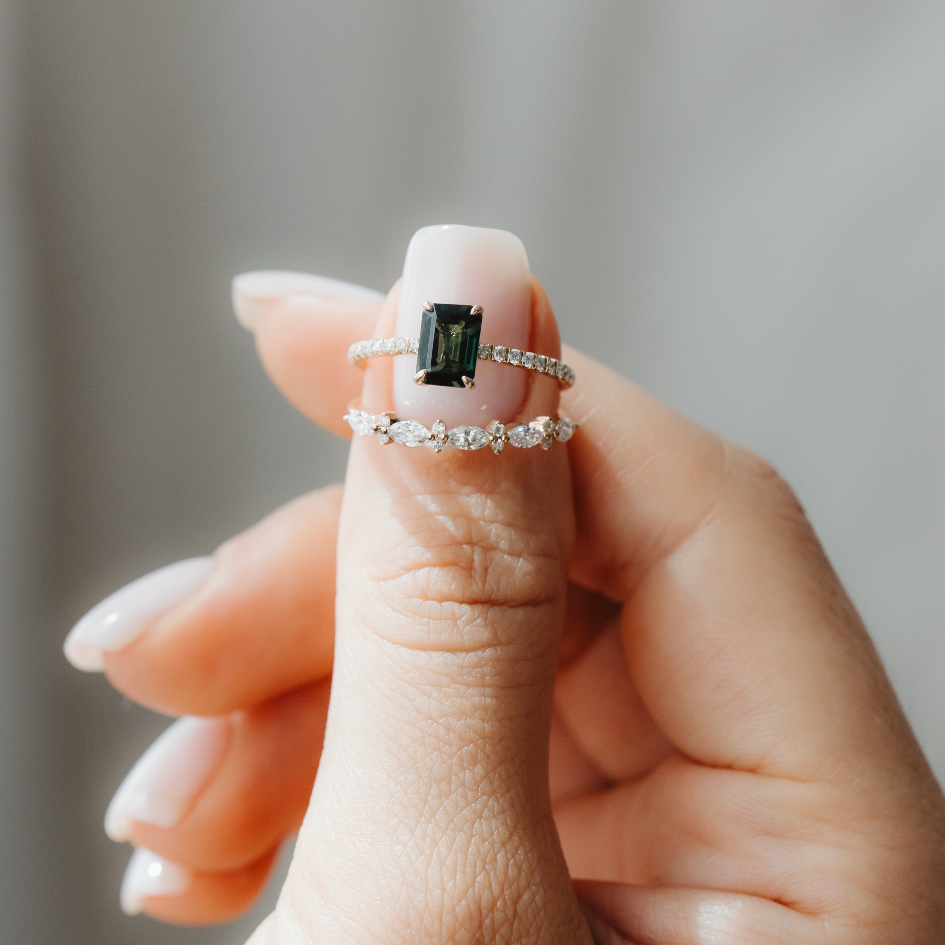 Gros plan d’une main tenant deux bagues en or jaune : une bague Elysian en saphir vert taille émeraude avec un anneau pavé de diamants, empilée au-dessus d’une alliance Fiorelle sertie de diamants taille marquise, sur un fond gris doux.