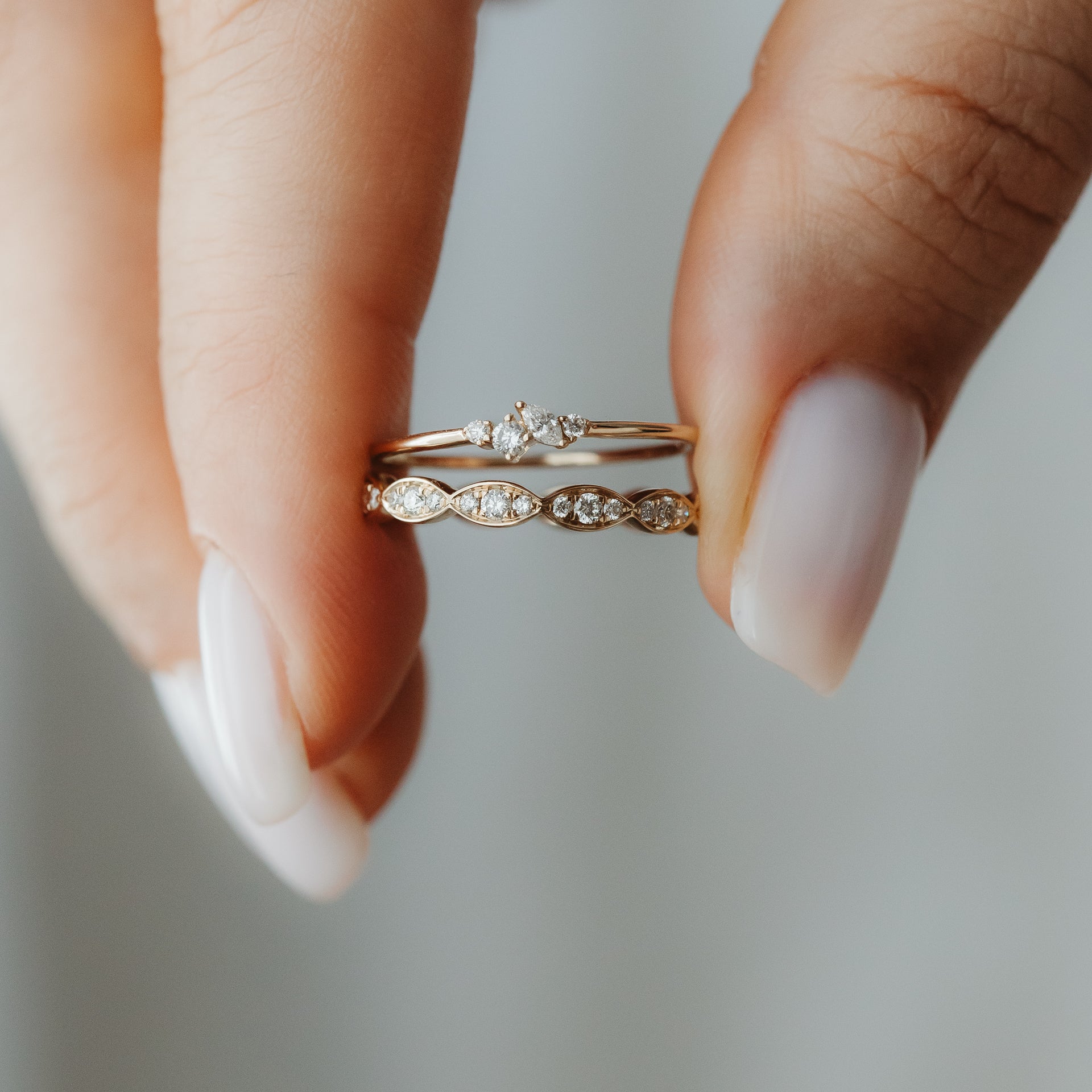 Gros plan de deux bagues à superposer en or jaune 14K serties de diamants, tenues entre les doigts — l'une avec un petit motif en grappe et l'autre avec des motifs en forme de marquise.