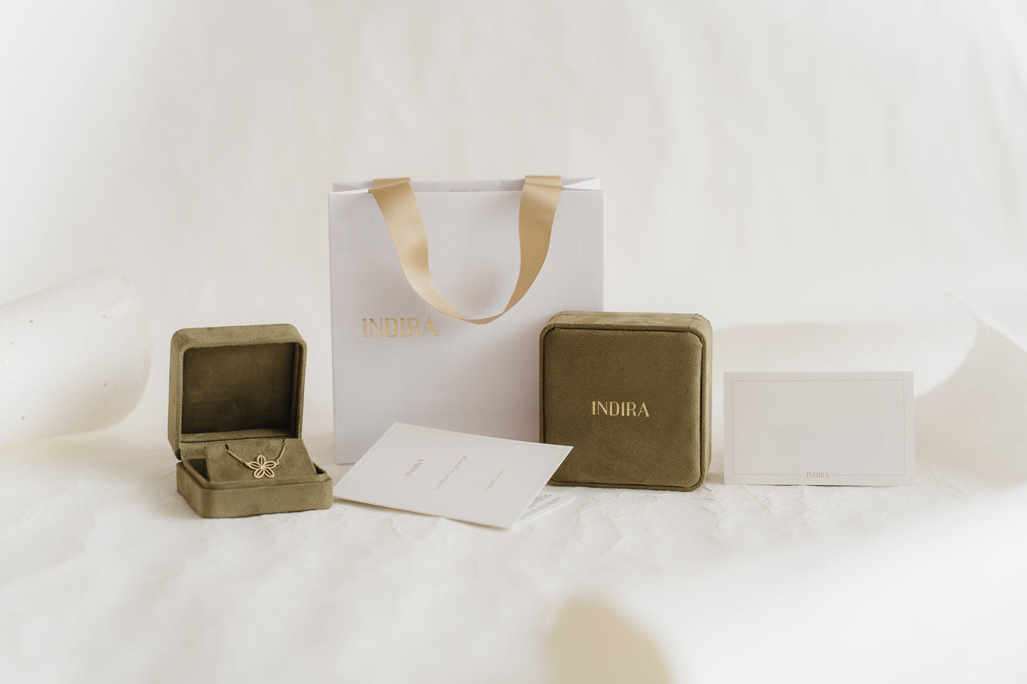 Ensemble d’emballage cadeau Indira : sac shopping blanc avec poignées en ruban doré, deux boîtes à bijoux en velours olive (l’une ouverte révélant un pendentif fleur en or) et cartes de marque sur un fond de studio blanc doux.