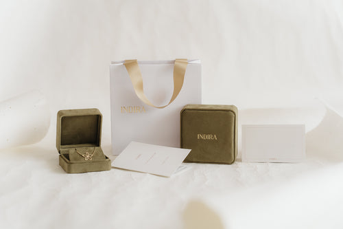 Ensemble d’emballage cadeau Indira : sac shopping blanc avec poignées en ruban doré, deux boîtes à bijoux en velours olive (l’une ouverte révélant un pendentif fleur en or) et cartes de marque sur un fond de studio blanc doux.