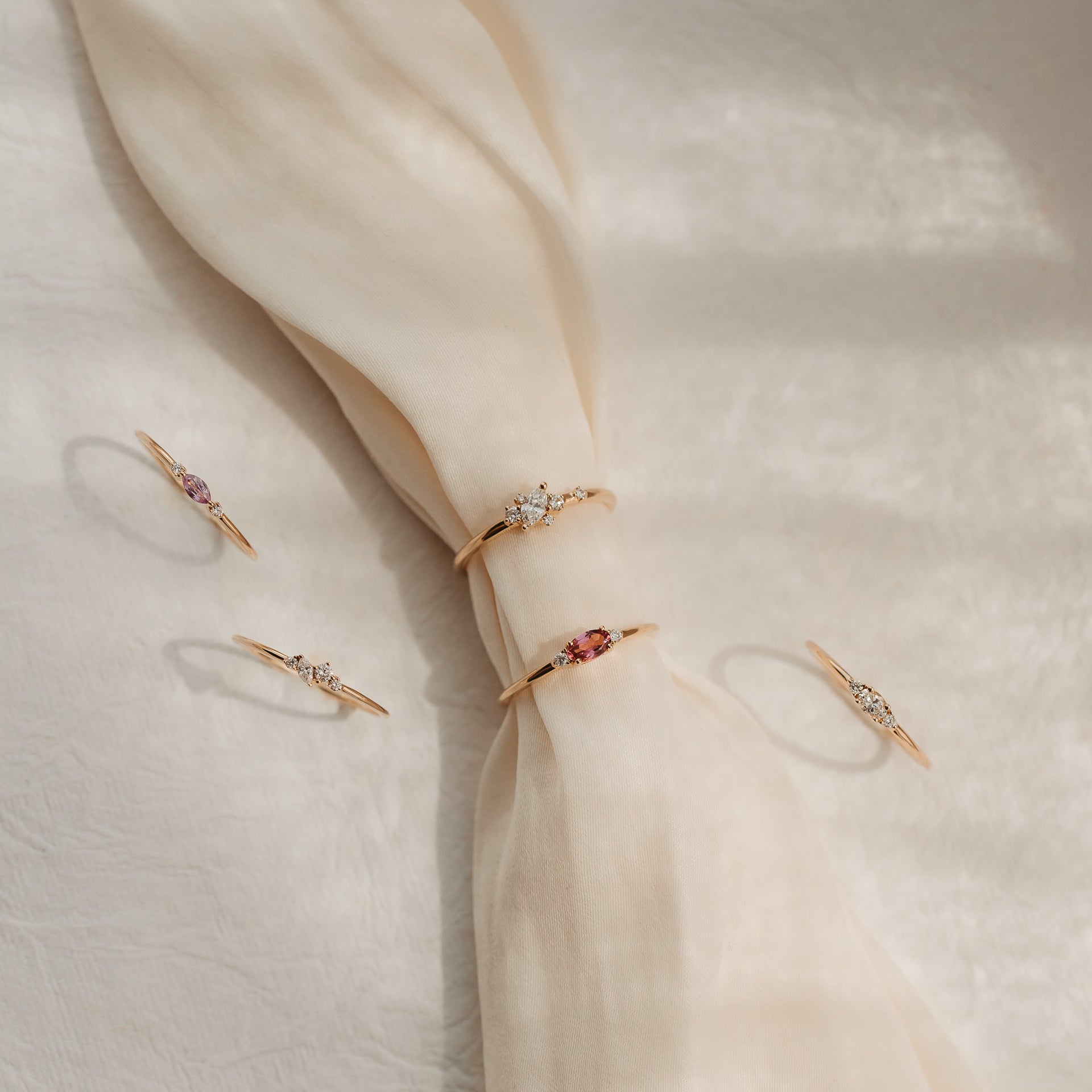 Cinq bagues délicates en or jaune 14k serties de diamants et de gemmes roses, présentées sur un tissu de soie crème noué, à la lumière douce du soleil.
