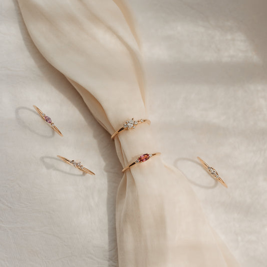 Cinq bagues délicates en or jaune 14k serties de diamants et de gemmes roses, présentées sur un tissu de soie crème noué, à la lumière douce du soleil.
