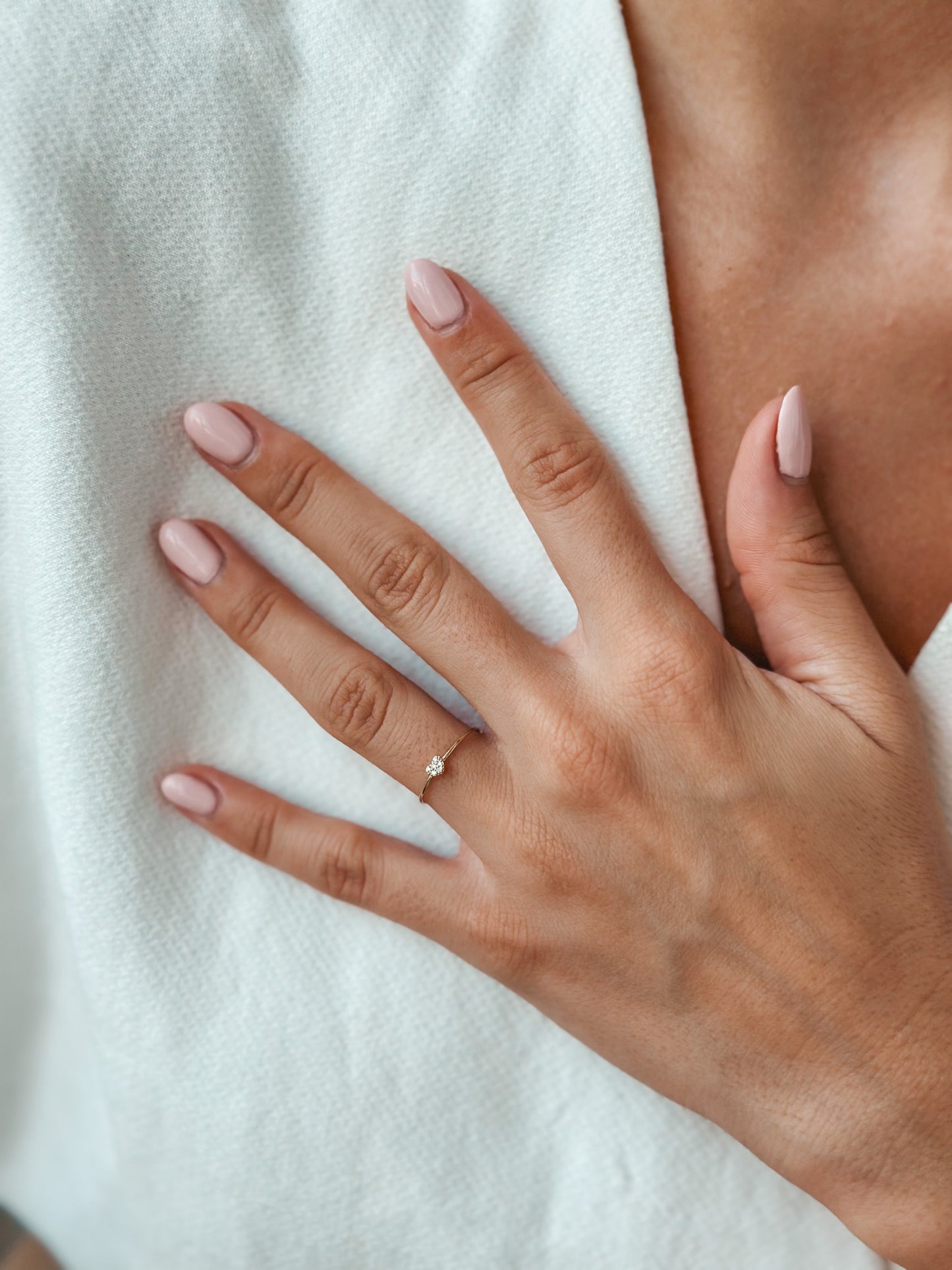 Gros plan d'une main aux ongles rose portant une bague en or délicate avec un petit diamant sur un fond de tissu blanc doux.