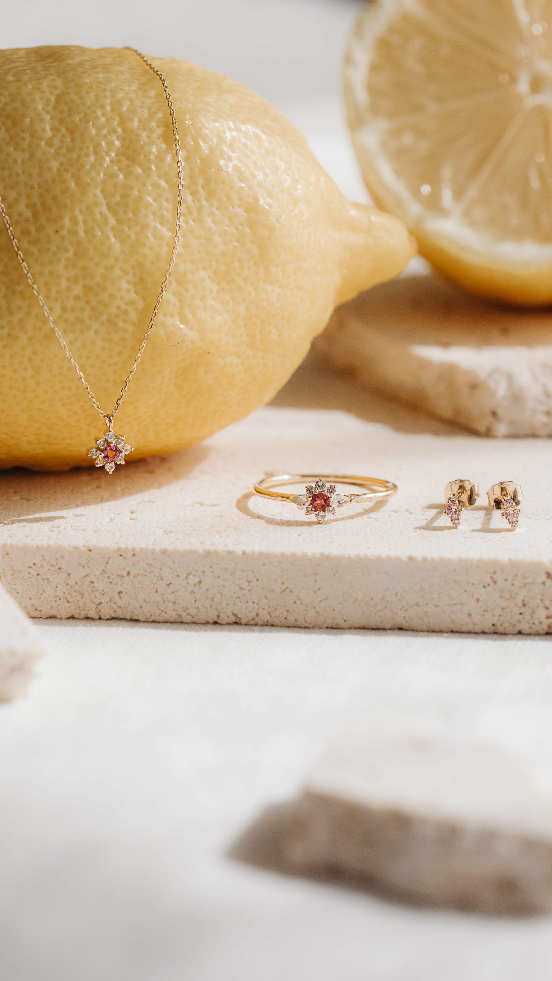 Clous d’oreilles Flora en or jaune 14K ornés de morganite rose et de saphirs blancs, présentés à côté d’une bague et d’un pendentif assortis, sur des carreaux en pierre avec des citrons.