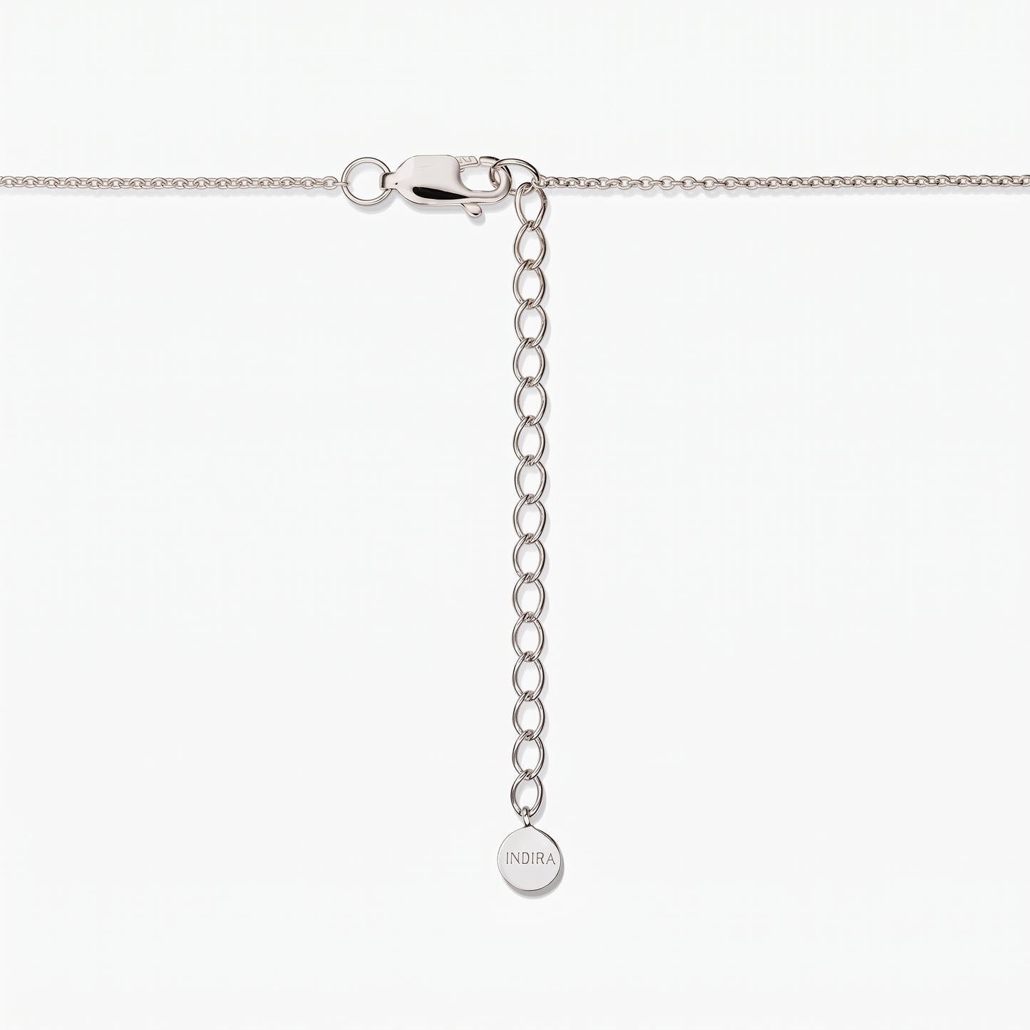 Gros plan sur un fermoir de collier en argent sterling avec une chaîne d’extension réglable et une petite plaque ronde gravée « INDIRA », sur fond blanc.