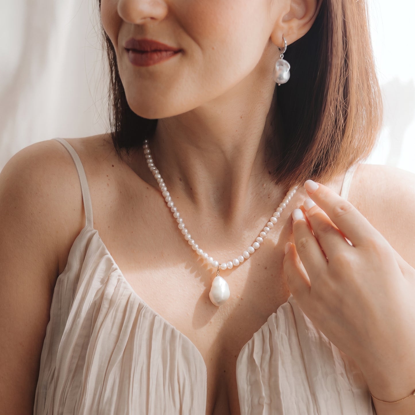 Gros plan d’un mannequin portant une boucle d’oreille pendante à perle baroque en argent 925 plaqué rhodium avec un petit détail en zircone, et un collier de perles assorti, la main près de la clavicule sous une douce lumière naturelle.