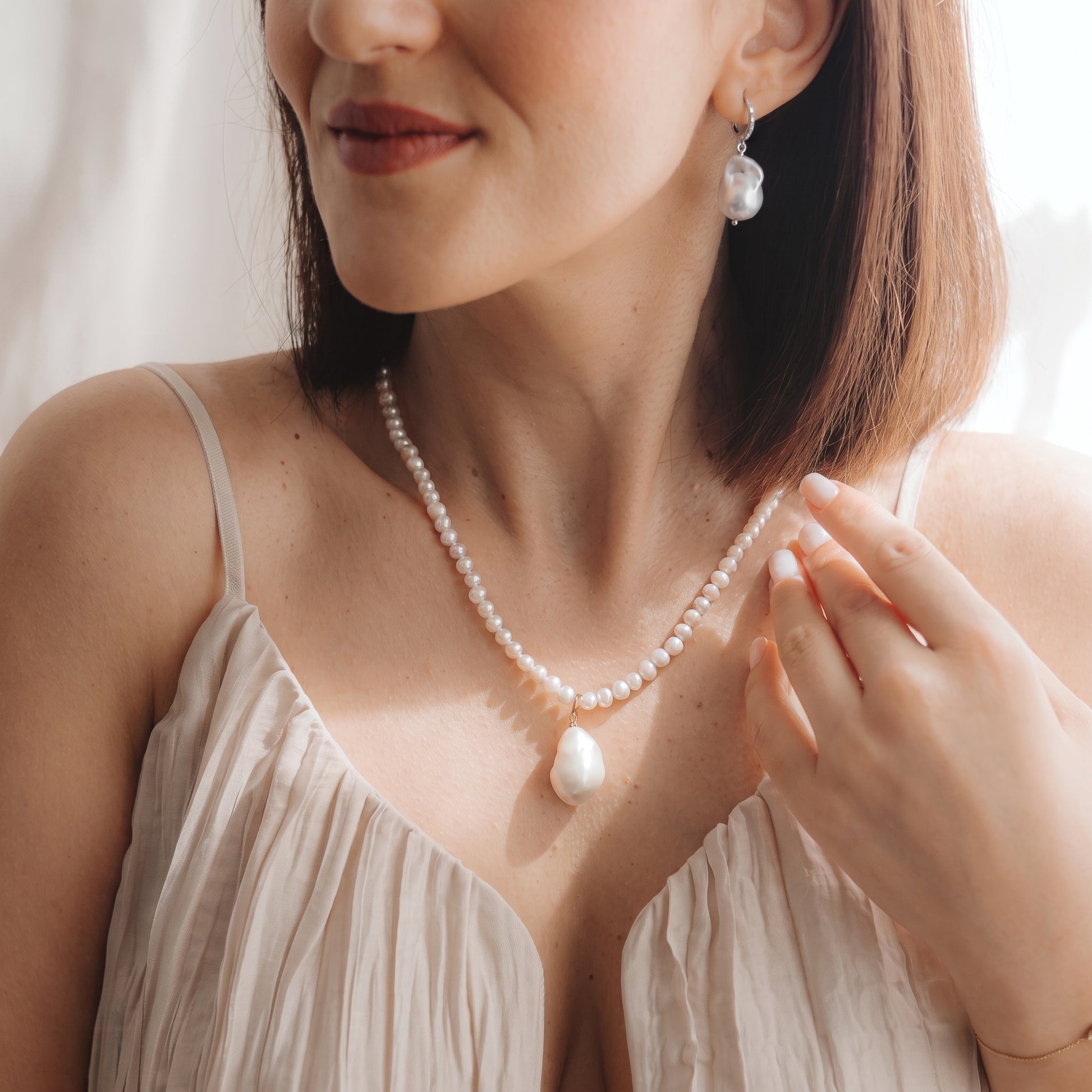 Gros plan d’un mannequin portant une boucle d’oreille pendante à perle baroque en argent 925 plaqué rhodium avec un petit détail en zircone, et un collier de perles assorti, la main près de la clavicule sous une douce lumière naturelle.
