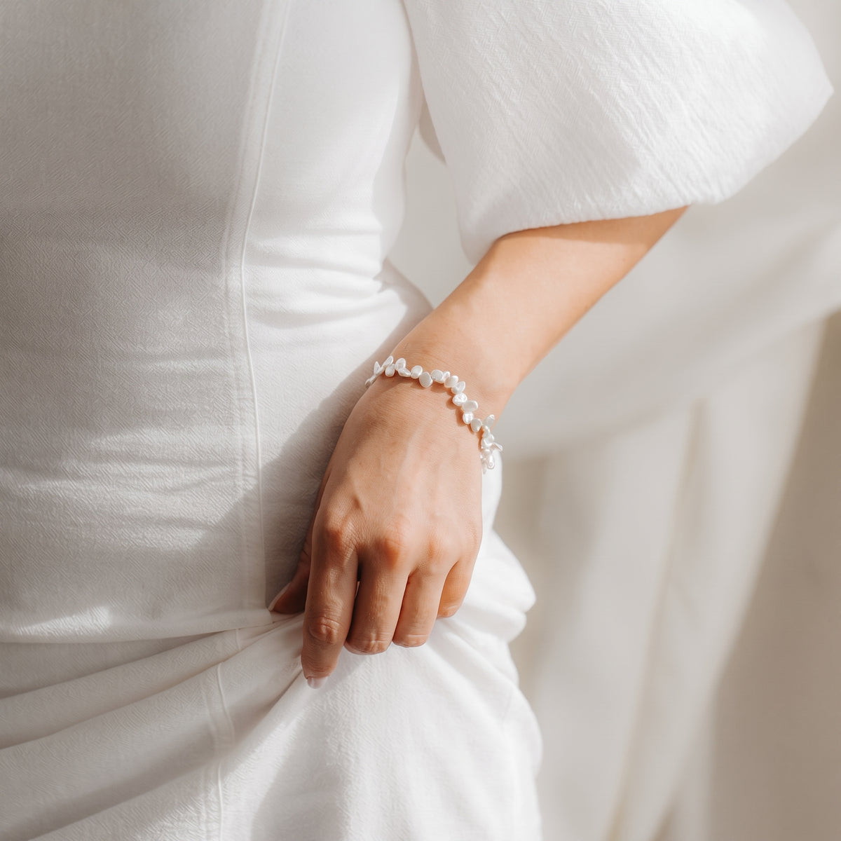 Gros plan d’un poignet orné d’un bracelet de perles Keshi en argent sterling plaqué or, porté avec une robe blanche sous une douce lumière naturelle.